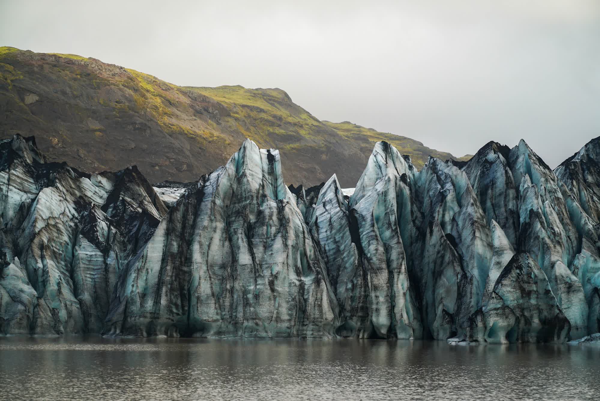 A wall of glaciers with mountains covered in moss in the backgrounand a lake in the foreground.