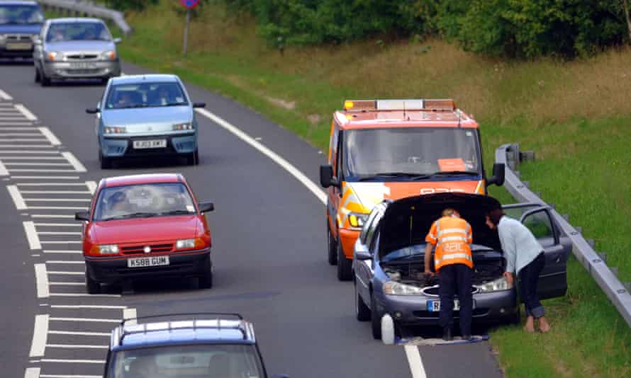 A car with its bonnet up and two people peering in, pulled partly off a busy road and onto a grass verge, with an orange RAC van behind it