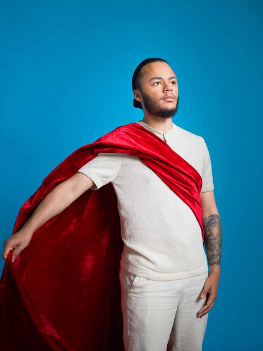 Kwajo Tweneboa, wearing a white T-shirt and trousers, and draped with red velvet, standing in a superhero pose