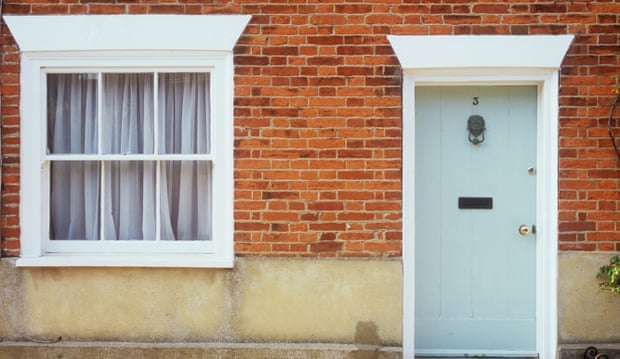 Simple wooden front door painted pale grey with white doorframe and lion head knocker and sash window with net curtains.