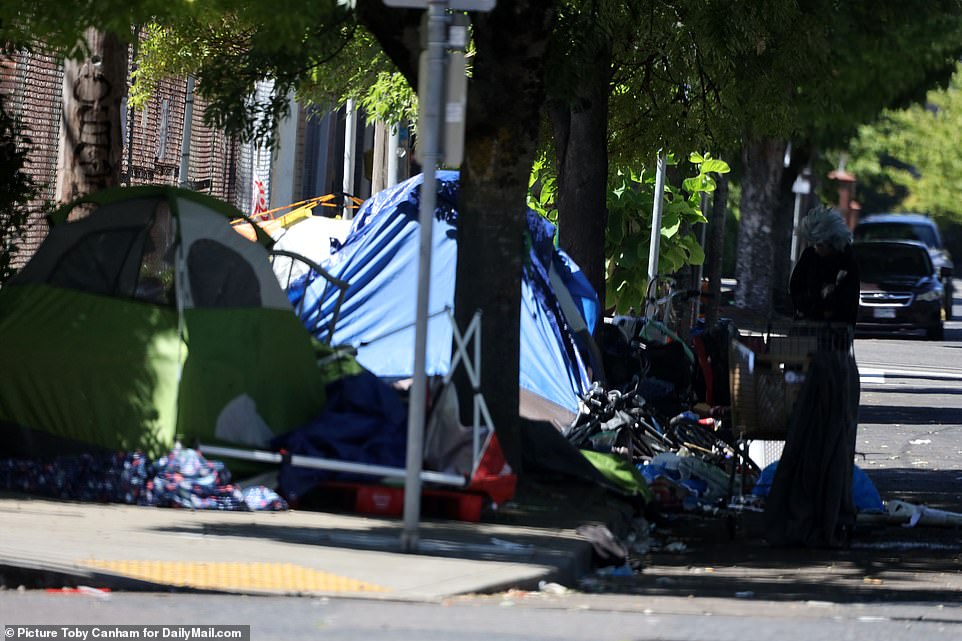 Tents are lined up along a residential street in Portland, with belongings strewn about and piles of trash in the roadway