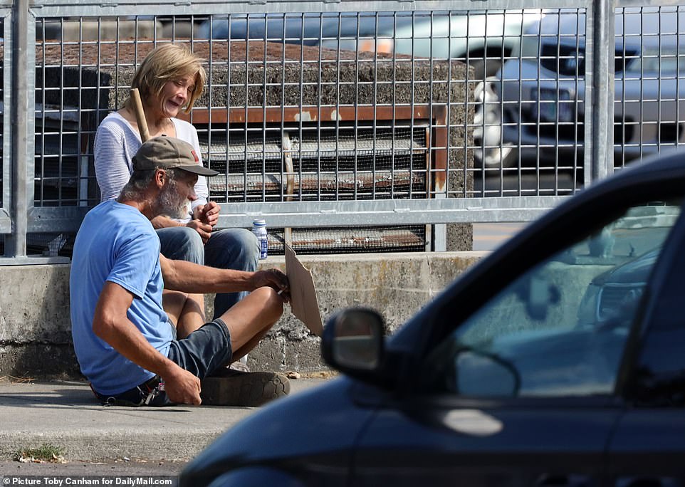 Two homeless people sit on the curb with a sign as they ask for money by passersby