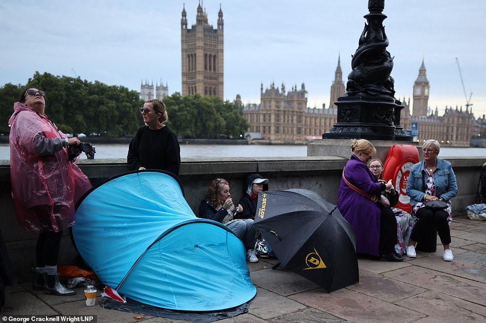 Two women, one in a waterproof poncho, stand next to a small tent along the bank of the River Thames in the queue