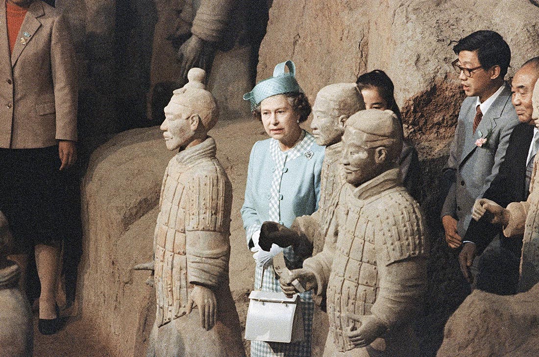 Queen Elizabeth stands among lifesize terracotta soldiers followed by Chinese dignitaries. The Queen wears a sky-blue pillbox hat with a sheer veil and a matching sky-blue suit jacket and dress with white and blue gingham edges.