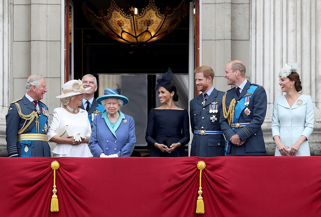 Queen Elizabeth standing on the balcony at Buckingham Palace surrounded by senior members of the royal family including Charles, Camilla, Meghan and Harry who are all looking at her smiling. The Queen wears a royal blue and teal coat and matching hat with black feathers.