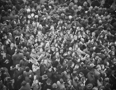 Mourners gather to catch a glimpse of the funeral procession. Some hold mirrors up in the air to try to get a view over the heads of others.