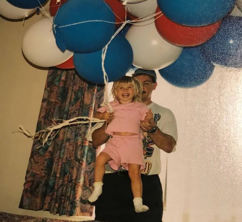 A man holds up a smiling girl as she holds onto some balloons. 