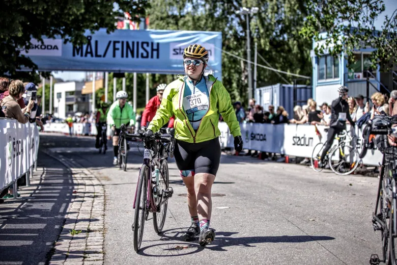 A woman wearing a helmet walks with her bike under a banner for the finish line.  