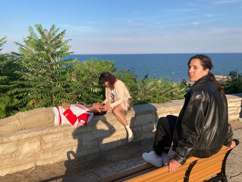 A man in a red and white jersey rests on a brick wall overlooking the ocean while a woman in a fringed jacket touches his head. A woman in a black leather jacket sits on the back of bench in front of them, looking over her shoulder. 