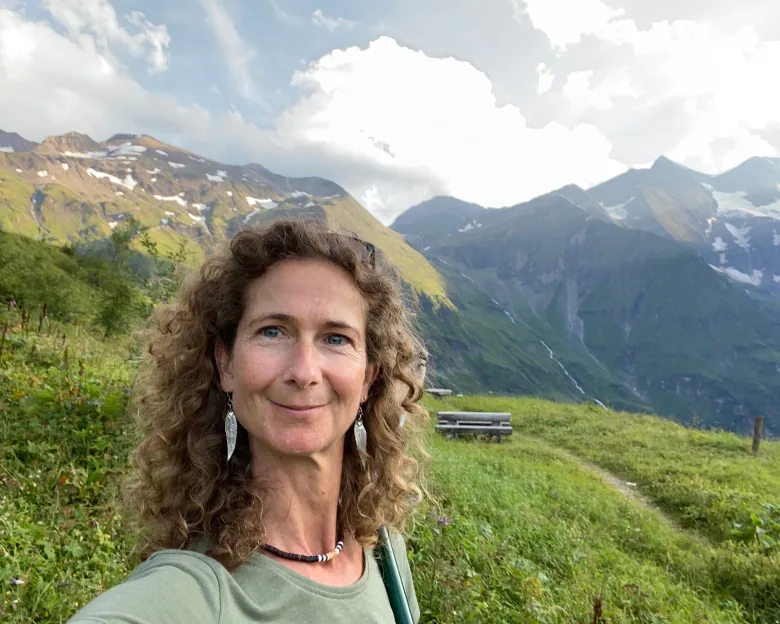 A woman with long, curly, light-blonde hair snaps a selfie in front of green fields and snowcapped mountains. 