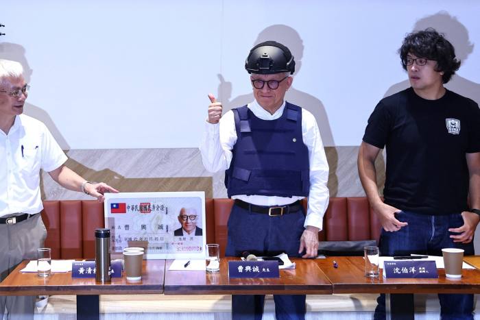 A man in a helmet and bulletproof vest stands at a desk at a press conference, giving the thumbs-up sign. Two men stand alongside him