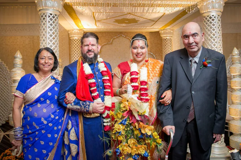 A bride and groom in traditional Indian clothing are flanked by an older woman and man holding a cane.  