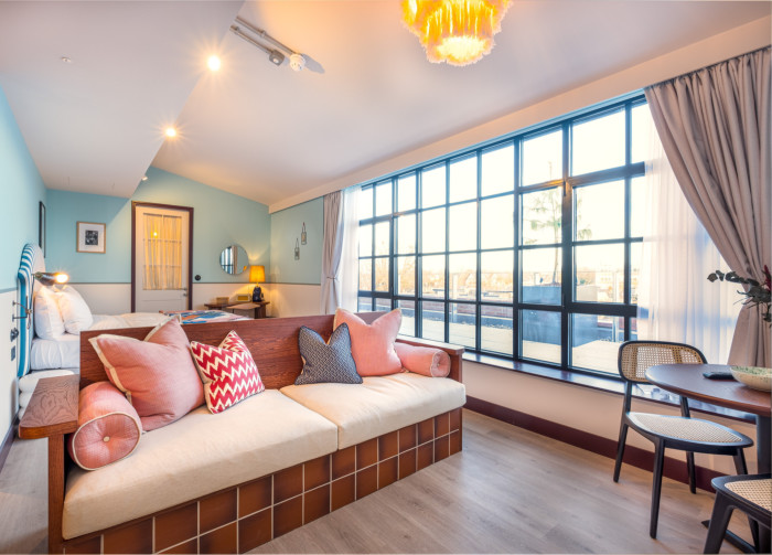 A sofa in front of a bed in a room in London’s room2 hotel, with pale-blue walls, a wooden floor and a large long window