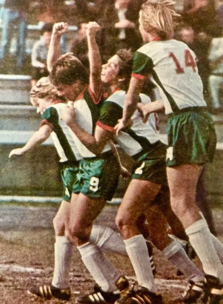 Four men in soccer uniforms celebrate on a soccer field. 