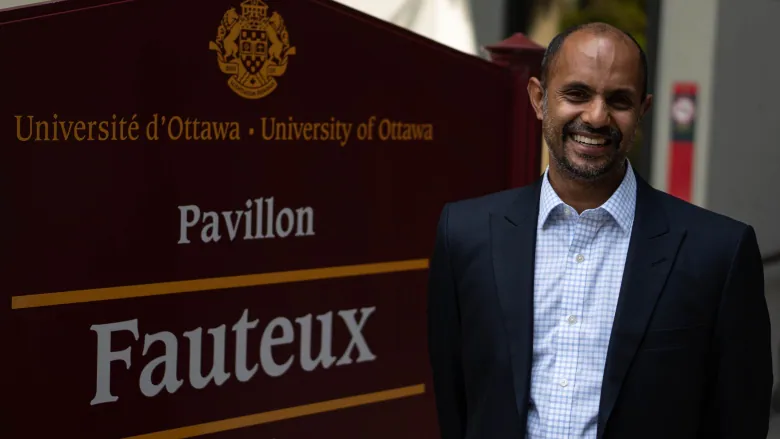 A man in a blue suit jacket smiles while standing next to a burgundy sign that says University of Ottawa.