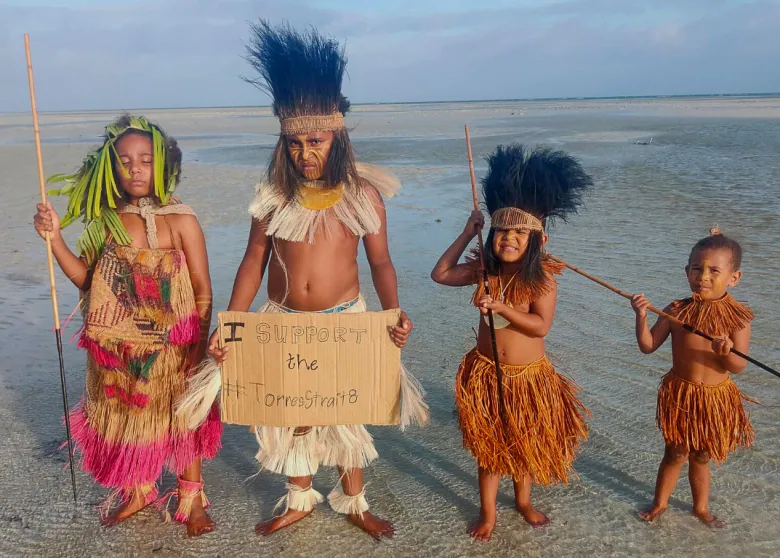 Four small children stand on a beach wearing traditional grass outfits. Three are holding tall sticks, and one is holding up a cardboard sign that reads: "I support the #TorresStrait8"