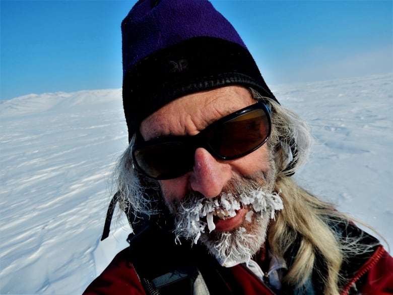 A selfie of a man with long gray hair and a snow-crusted mustache and bearded. He's wearing sunglasses and a toque. The backdrop is clear white snow and bright blue sky.