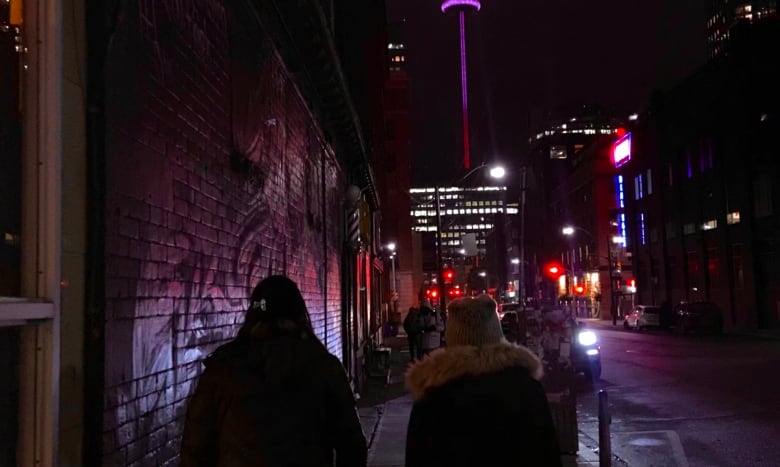 Two women walk on a sidewalk. The CN Tower is lit up at nighttime ahead of them. 