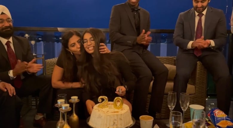 A woman kisses her daughter on the cheek as others cheer on. A cake with the number ‘20’ is on the table in front of them.    