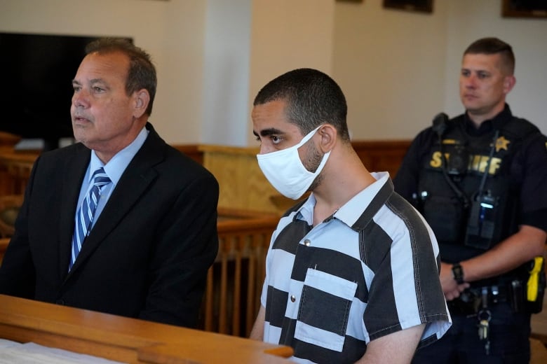 A man in a COVID-19 mask and a prison-issued shirt listens in a court session.