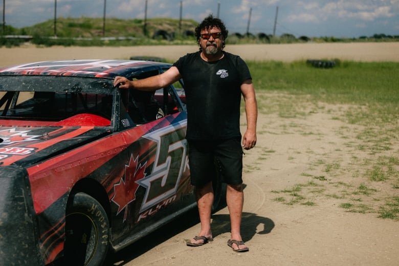 A man stands next to a racecar with his hand on the frame. The car is black and red with a large number five, a maple leaf and the name "Klym" where the door would typically be.