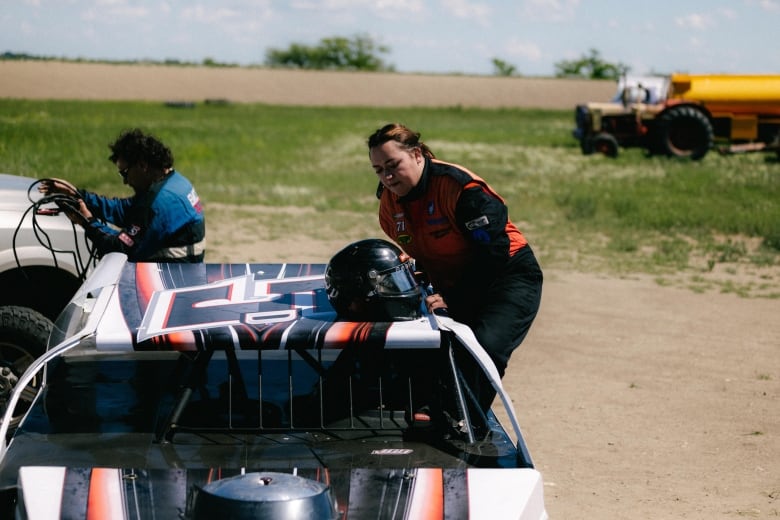 A woman wearing a racing suit lowers herself into the driver's seat of a car through the window. Her helmet rests on the roof.