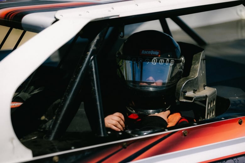 A woman sits in the driver's seat of a racecar wearing a helmt and racing suit.