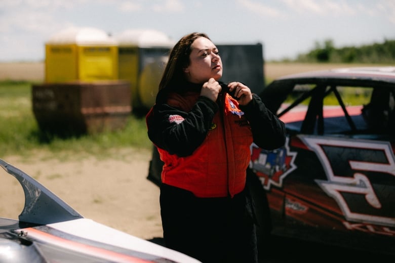 A young woman does up her orange and black racing suit as she stands between two racecars.