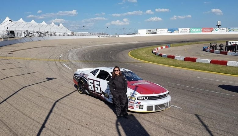 A young woman in a racing suit stands in front of a white and red racecar on an oval track.