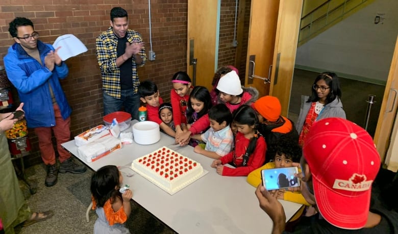 A group of kids crowd around a table to cut a white cake with strawberry decorative frosting. 