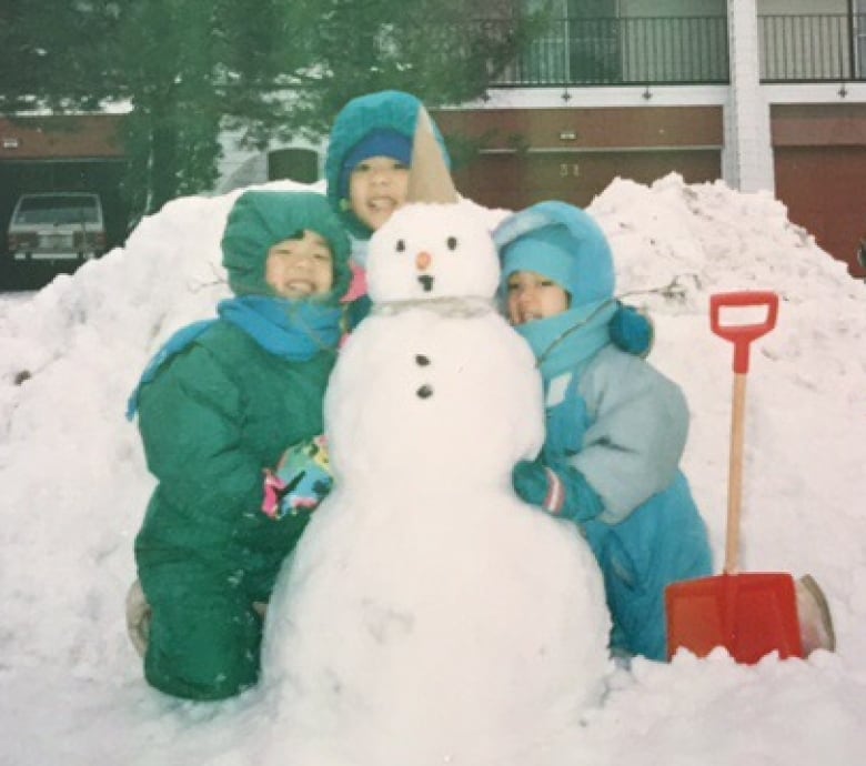 Three girls in snowsuits hug a snowman.