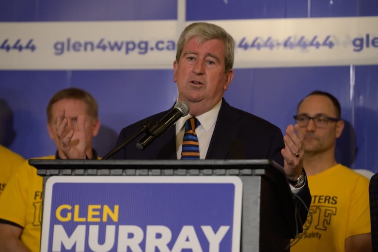 A man is standing at a podium speaking into a microphone, with a sign that says "Glen Murray."