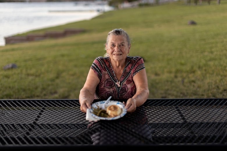 A smiling elderly woman sits at a picnic table holding up a plate of food.