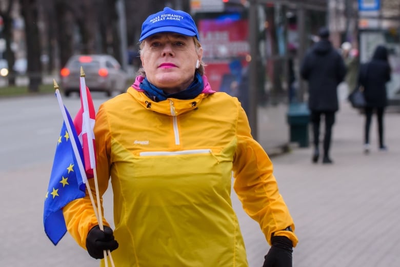 A woman in a yellow jacket and a blue ballcap that reads "Make Humanity Great Again" runs in the street while holding two flags, one for Latvia and one for the European Union.