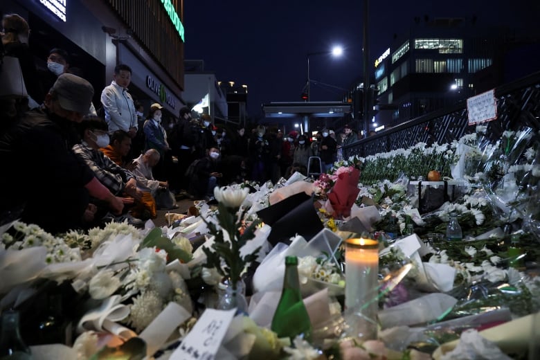 Dozens of people stand on the side of a street at night next to giant piles of flowers and candles.