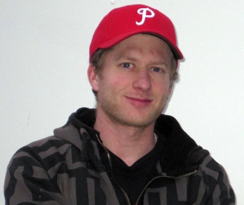 A young white man with a red baseball cap and grey and black patterned hoodie is giving slight smile in front of a white background.