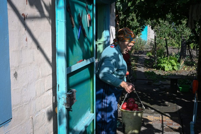 An old woman in headscarf emerges from her turquoise-painted door with a bucket of red apples