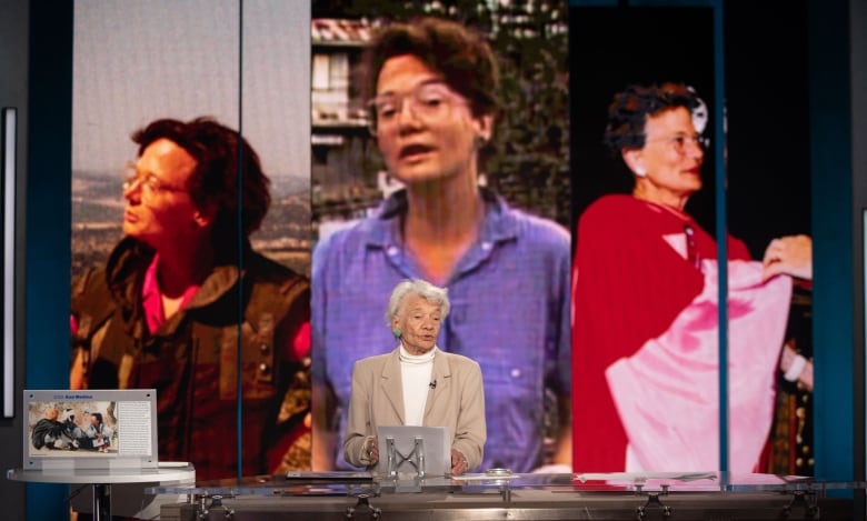 An elderly woman stands, midspeech, in front of a desk. Behind her are three larger than life photos, all of her as a younger woman. On the right a woman wearing a flak-jacket looks out of frame. In the centre a woman looks toward the camera, midspeech. On the right a woman draped in a billowing red robe looks off camera.