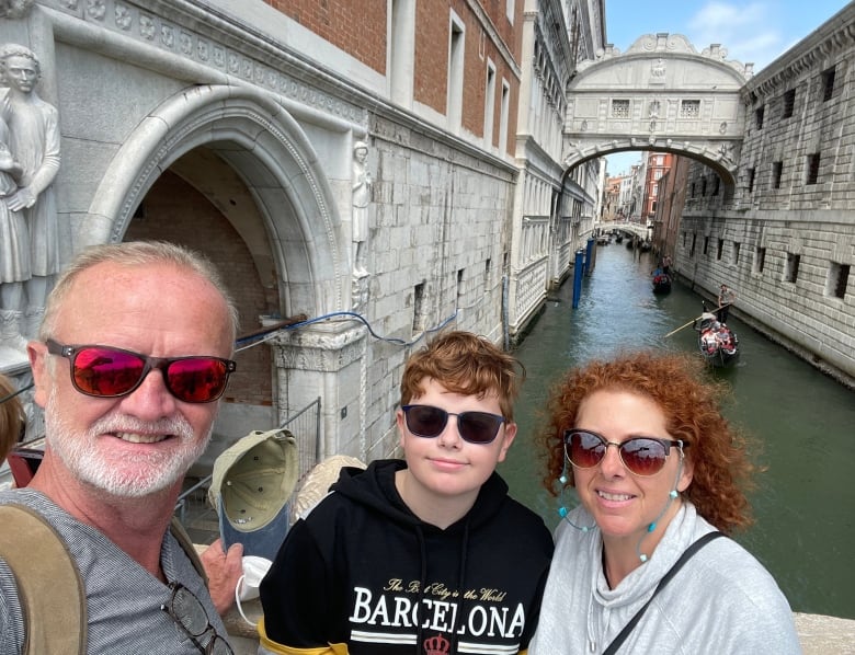 A smiling family of three -- a dad, teenage son and mom -- take a selfie next to a canal in Venice, with stone architecture and gondolas in the background.