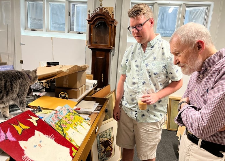 Two men look at a number of paintings propped up on a table.