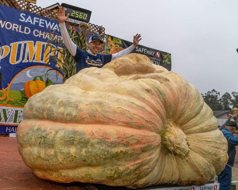 A man stands on a stage behind a massive pumpkin the size of a dumpster, holding his hands above his head.