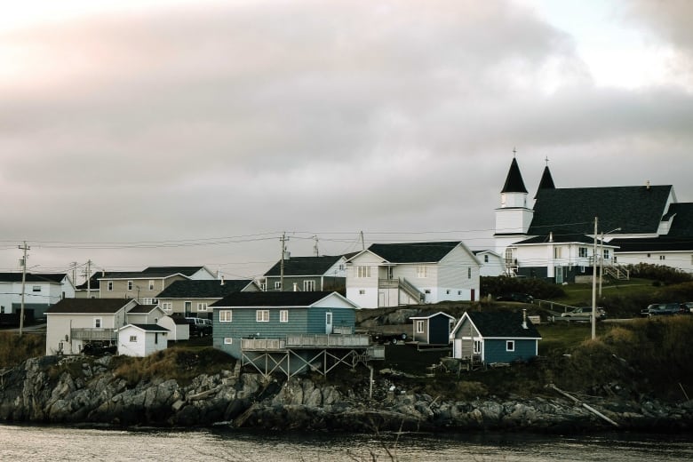 A house clings to rocks by the sea with a church in the background