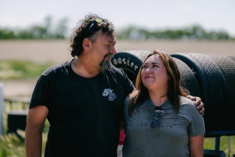 A middle-aged man and young woman stand in front of a rack of tires, staring affectionely at one another with smiles on their faces.