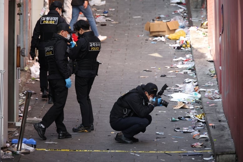 A uniformed police officer kneels and snaps photos of the debris lining the side of an alley as three other officers inspect the scene. 