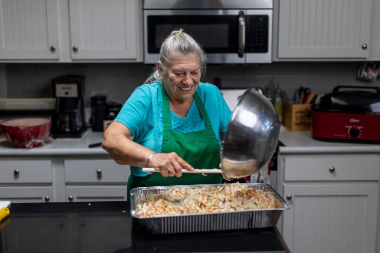 An elderly woman in an apron smiles as she stands in a kitchen scooping ingredients from a bowl into an aluminum container. 