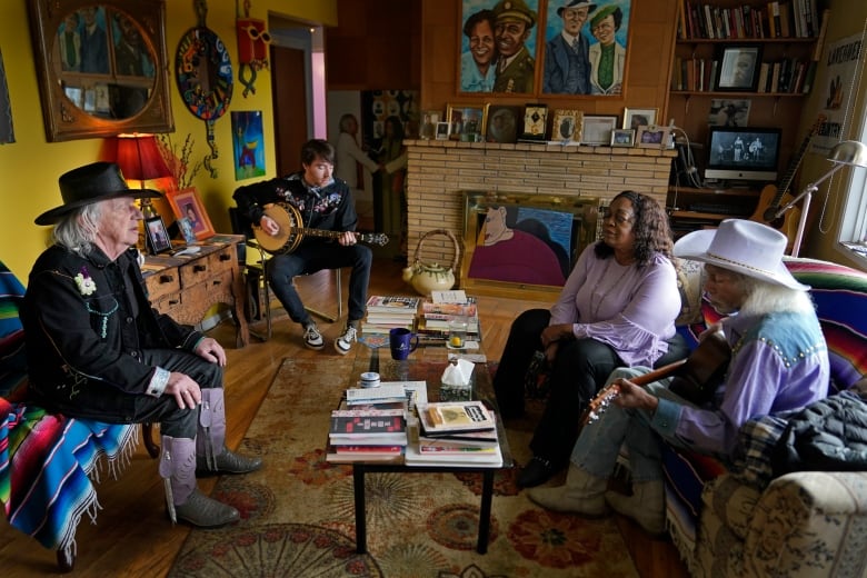 Three men and a woman sit around a living room with yellow walls and colourful art filled with books and other ephemera. One of the men is strumming a banjo, and another is holding a guitar. Two of the men are wearing cowboy hats.