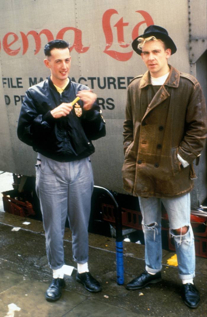 Two young men stand next to each other on a street wearing worn jeans. One man has a black bomber jacket, the other a worn leather jacket and hat