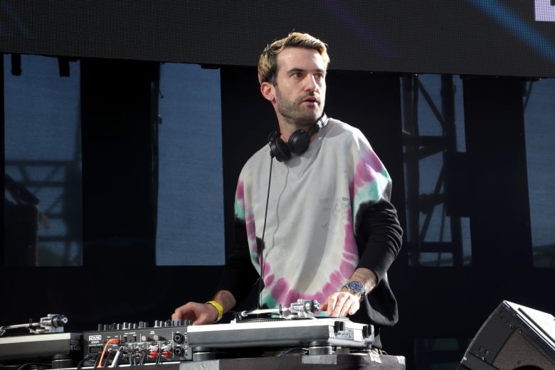 A man with died blonde hair, a five o'clock shadow and a tie-dye sweatshirt looks to one side as he DJs on stage. 