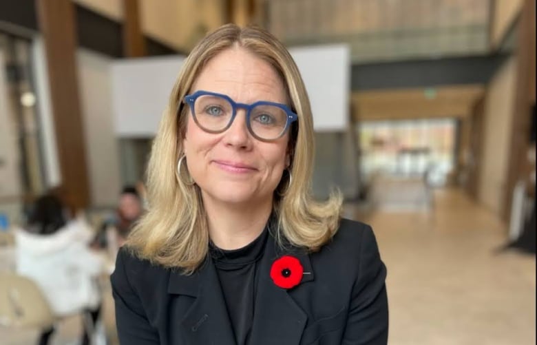 A woman wearing blue glasses, a black blazer and a Remembrance Day poppy smiles while sitting in a large, bright indoor atrium.