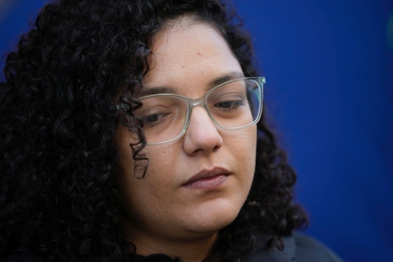 A close up on the face of a frowning woman with curly black hair and clear glasses looking downward. 